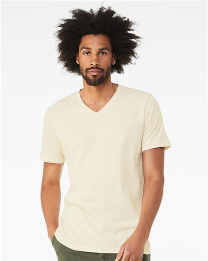 A man with medium brown skin and curly afro hair wears the BELLA + CANVAS 3005 unisex V-neck in light beige, made from Airlume combed cotton, and dark green pants. He stands against a white background with a neutral expression.