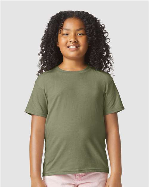 Smiling child with curly hair wears a Gildan Youth 64000B CVC t-shirt in olive green and light pink pants, standing against a plain light gray background.