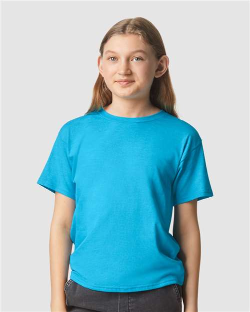 A young person with long brown hair, wearing a bright blue Gildan Youth 64000B CVC t-shirt and dark pants, stands against a plain light gray background with a slight smile.