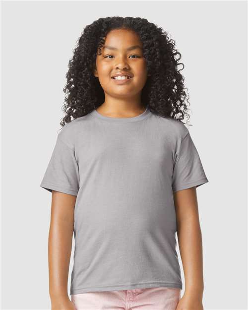 A smiling child with long curly hair wears a Gildan Youth 64000B CVC t-shirt in plain gray and light pink pants, standing against a light gray background. The shirt is made from OEKO-TEX certified Softstyle cotton by Gildan.