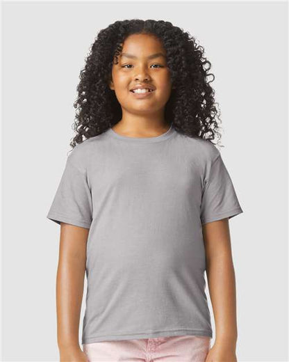 A smiling child with long curly hair wears a Gildan Youth 64000B CVC t-shirt in plain gray and light pink pants, standing against a light gray background. The shirt is made from OEKO-TEX certified Softstyle cotton by Gildan.