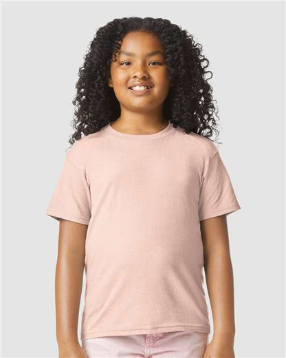A smiling child with long, curly hair wears a Gildan Youth 64000B CVC t-shirt by Gildan, OEKO-TEX certified for safety and comfort, standing against a plain gray background.