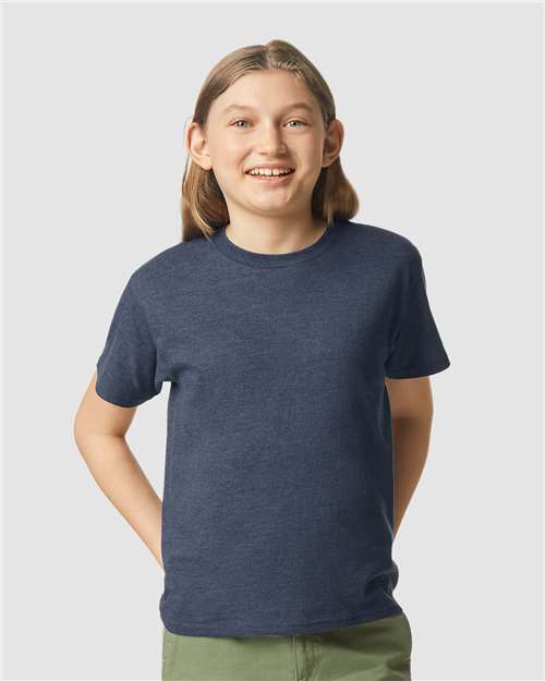 Smiling child with long light brown hair in a navy blue Gildan Youth 64000B CVC t-shirt by Gildan and green pants, standing against a plain light gray background.