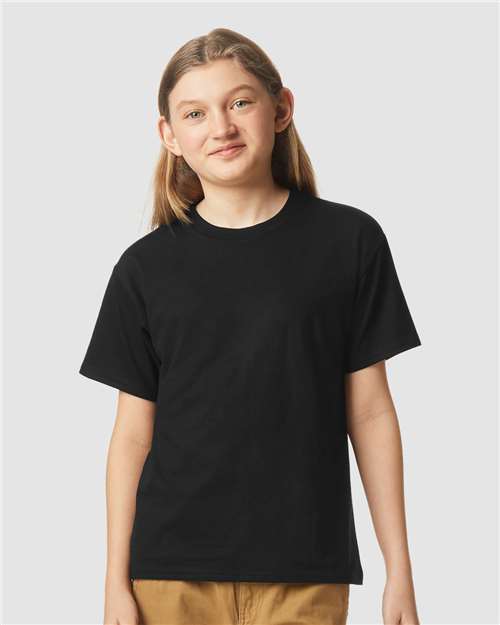 A young person with long, light brown hair wears a plain black Gildan Youth 64000B CVC t-shirt and tan pants, standing against a light gray background and smiling slightly at the camera.