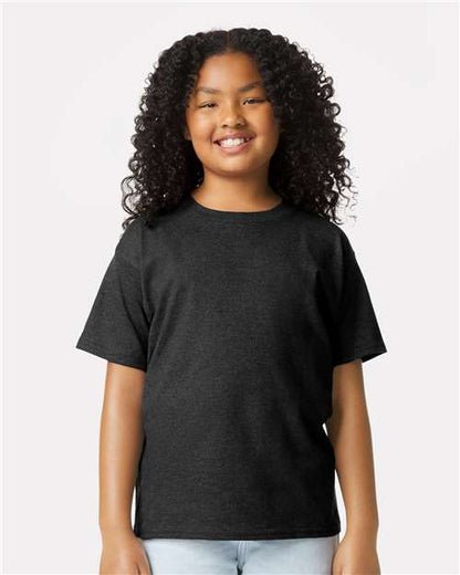 A smiling child with curly hair wears a plain black Gildan Youth 64000B CVC t-shirt and light blue jeans, standing against a simple light-colored background.