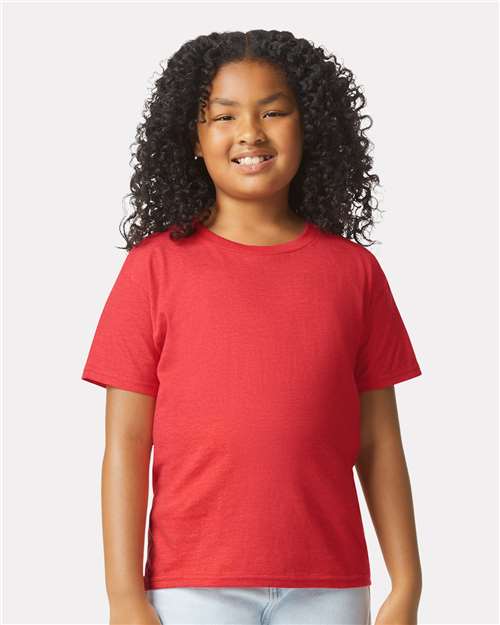 A cheerful child with curly hair wears a bright red Gildan Youth 64000B CVC t-shirt by Gildan, made with sustainable practices, and light blue jeans, standing against a plain light background.