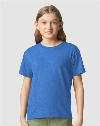 A young person with long brown hair stands against a plain light gray background, wearing a bright blue Gildan Youth 64000B CVC t-shirt and olive green pants, facing forward with a neutral expression.