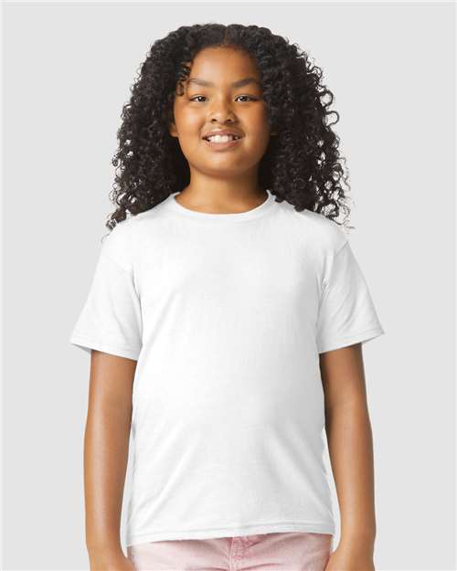 Smiling child with long curly hair wearing a Gildan Youth 64000B CVC plain white t-shirt and light pink pants, standing against a plain light gray background.