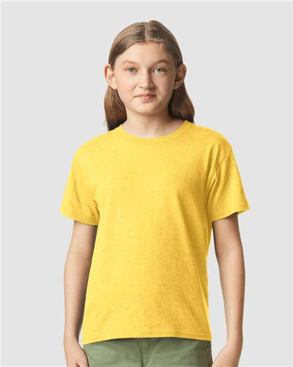 A young person with long brown hair stands against a light gray background, wearing a yellow Gildan Youth 64000B CVC t-shirt and green pants, looking forward with a slight smile.