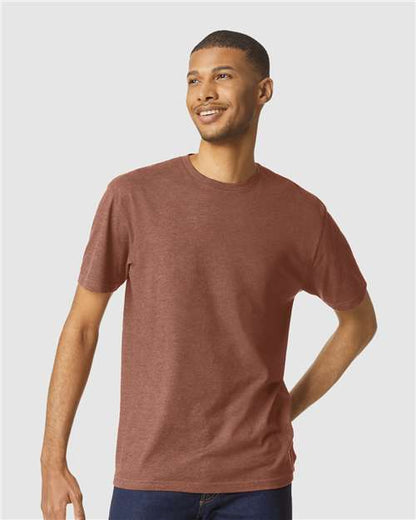 A smiling man wears a Gildan Softstyle 64000CVC heathered reddish-brown t-shirt made from ring-spun cotton and dark jeans, standing against a plain light gray background.
