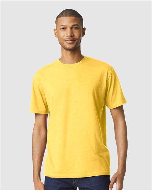 A man stands facing forward, wearing a bright yellow Gildan Softstyle 64000CVC T-shirt and dark jeans against a plain light gray background.