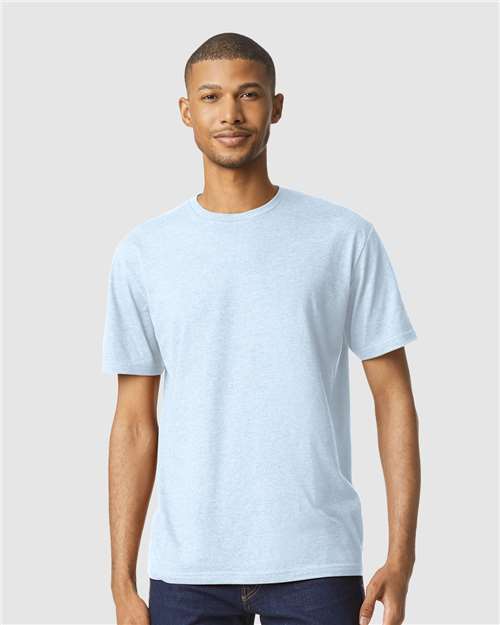 A man faces forward, wearing a Gildan Softstyle 64000CVC light blue T-shirt and dark jeans against a plain light gray background.