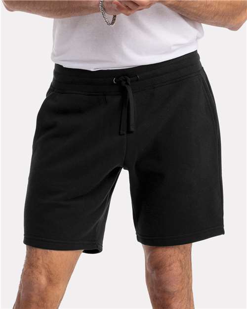 A person wears a white Next Level 9903 combed ringspun cotton T-shirt and black drawstring shorts, standing with arms crossed from waist to mid-thigh against a plain light background.