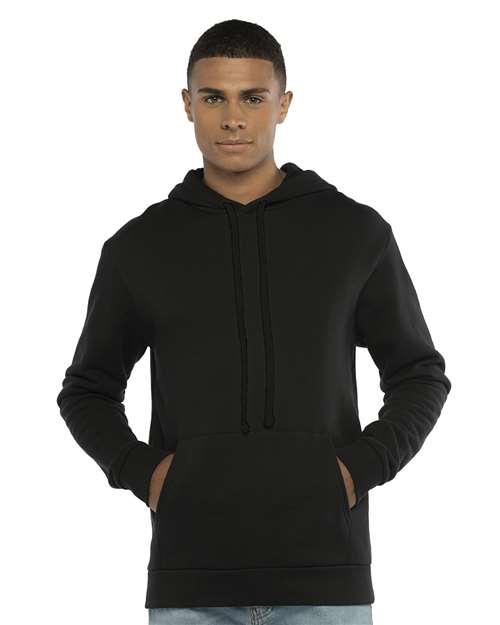 A person faces forward wearing the Next Level 9303 plain black cotton-polyester hoodie with a jersey-lined hood and front pocket, hands tucked in. The background is white.