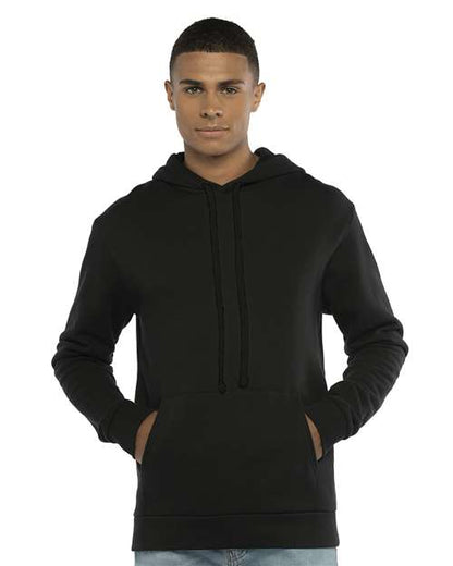 A person faces forward wearing the Next Level 9303 plain black cotton-polyester hoodie with a jersey-lined hood and front pocket, hands tucked in. The background is white.
