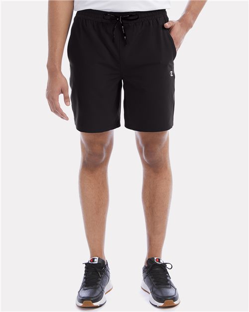 A person wears Champion CHP150 black polyester spandex shorts with an elastic waistband and black sneakers with white soles, standing against a plain light background. Only the lower torso and legs are visible.