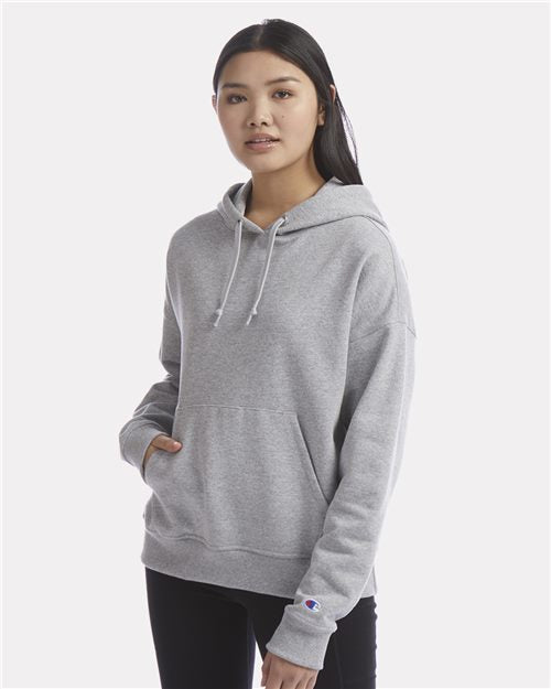 A young woman with long dark hair is wearing a Champion S760 light gray hoodie with a relaxed fit and front pocket, paired with black pants, standing against a plain white background.