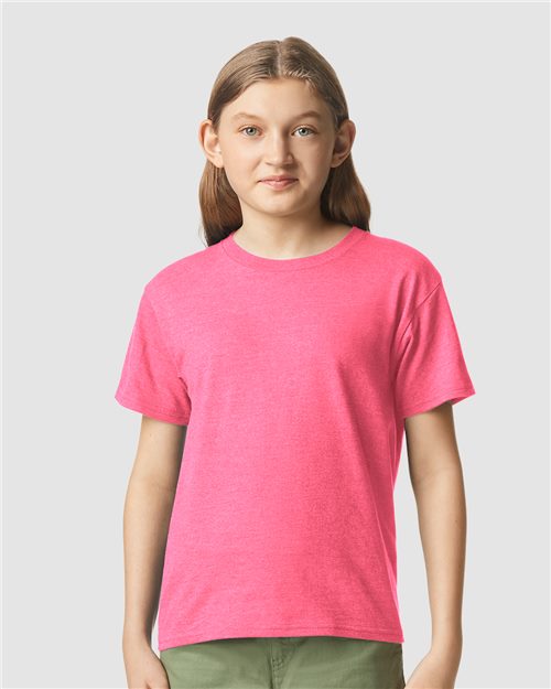 A young person with long light brown hair faces forward, wearing a bright pink Gildan Youth 64000B CVC t-shirt and green pants against a plain light gray background.