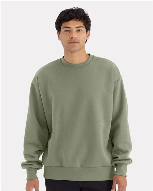 A person wearing the Next Level 9007 relaxed fit crewneck sweatshirt in sage green combed ring-spun cotton stands facing forward in dark pants against a plain light gray background.