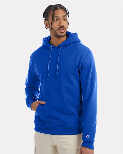 A man with braided hair wears the Champion S700 bright blue hoodie, made from recycled polyester, and beige pants, standing against a plain light background with one hand in his pocket and looking to the side.