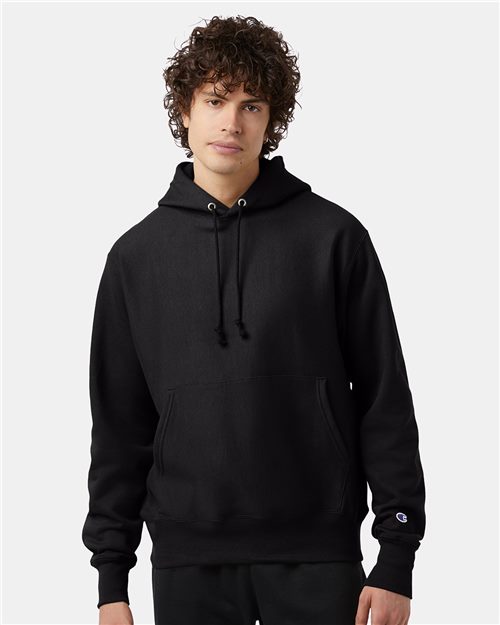 A young man with curly brown hair wears a black Champion S101 hoodie with a front pocket and drawstrings, standing against a plain light gray background.