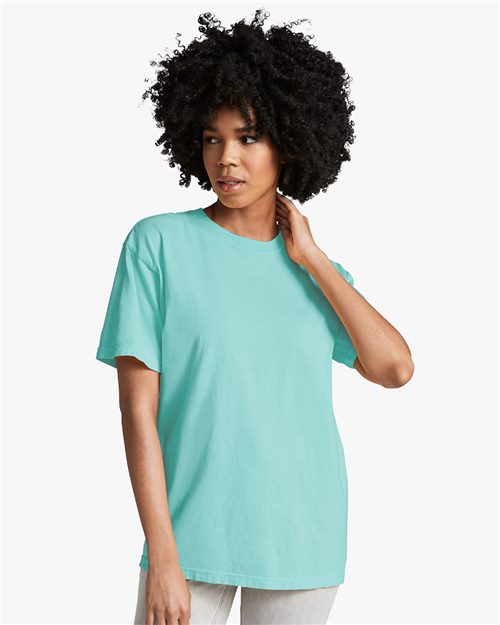 A woman with curly hair wears the loose-fitting, light blue Comfort Colors 1717 t-shirt, OEKO-TEX certified, and light-colored pants, standing against a plain white background and looking slightly to her left.