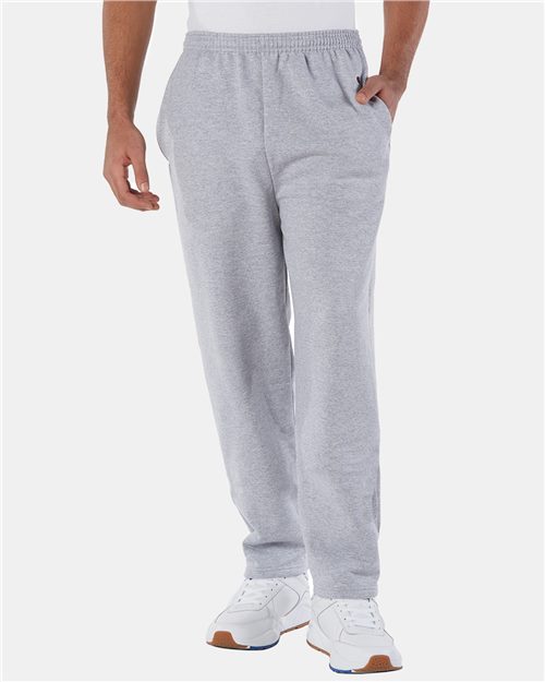 Someone stands against a plain white background wearing Champion P800 light gray sweatpants made with recycled polyester, hands in pockets, and white athletic shoes.