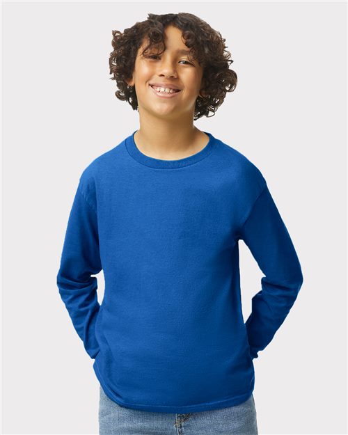 A smiling child with curly hair wears a bright blue Gildan 5400B long-sleeve shirt and jeans, standing with hands in pockets against a plain light gray background.