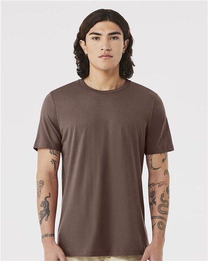 A person with long brown hair stands facing forward, wearing a brown BELLA + CANVAS 3001 unisex t-shirt made from Airlume combed and ring-spun cotton. Their tattooed arms rest at their sides against a plain white background.