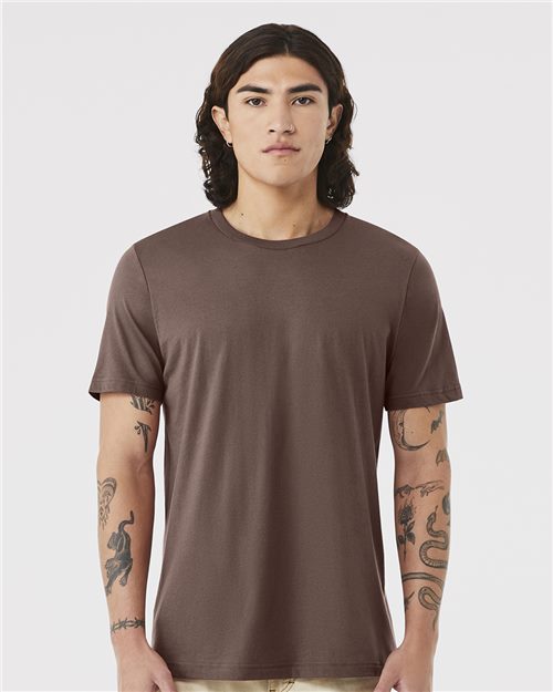 A person with long brown hair stands facing forward, wearing a brown BELLA + CANVAS 3001 unisex t-shirt made from Airlume combed and ring-spun cotton. Their tattooed arms rest at their sides against a plain white background.