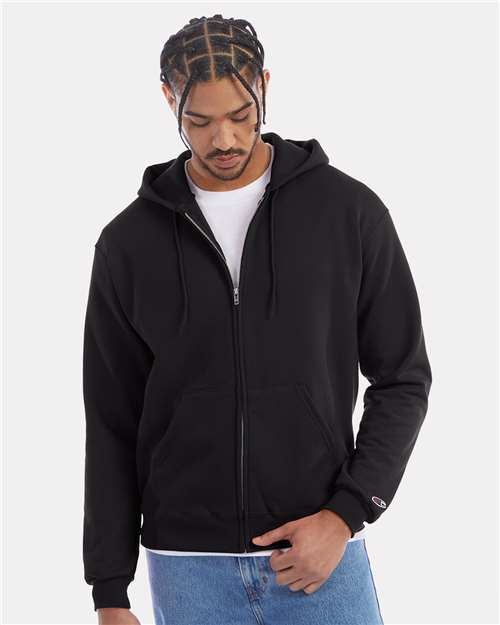 A man wearing a black Champion S800 hoodie made from recycled polyester over a white t-shirt and blue jeans stands against a plain light background, looking down while holding the edge of the hoodie.