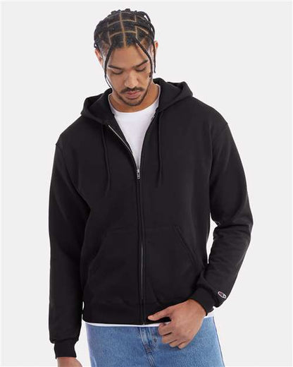 A man wearing a black Champion S800 hoodie made from recycled polyester over a white t-shirt and blue jeans stands against a plain light background, looking down while holding the edge of the hoodie.