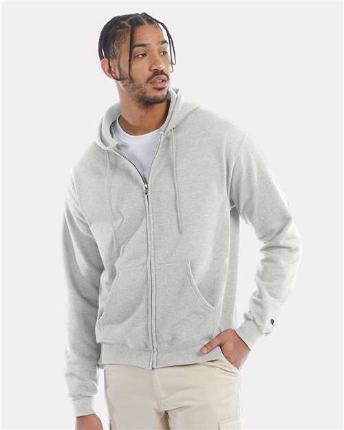 A man wears a light gray Champion S800 hoodie, made with recycled polyester, over a white tee and beige pants, standing against a plain light background and looking to his left.