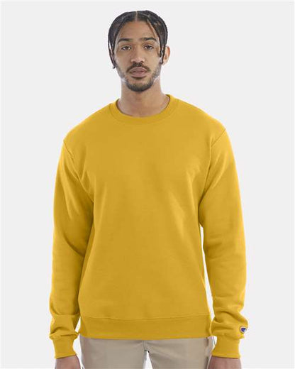 A man with braided hair wears a bright yellow Champion S600 sustainable sweatshirt made from recycled polyester and beige pants, standing against a plain light gray background and facing forward with a neutral expression.