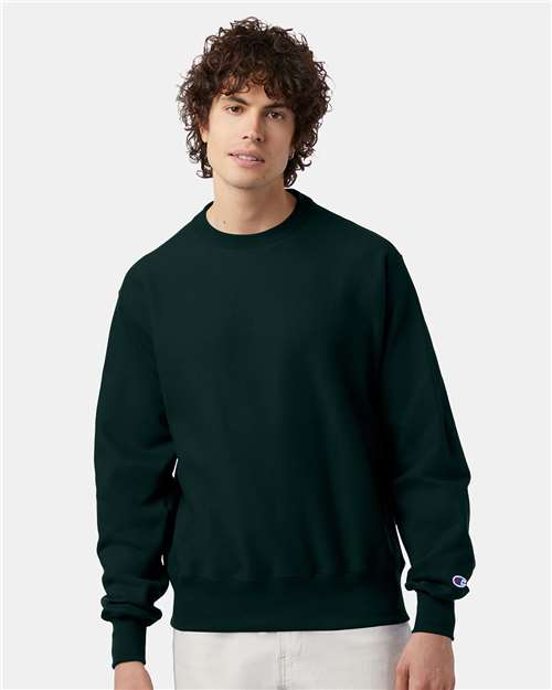 A young man with curly brown hair wears the Champion S149 sweatshirt and light pants, standing against a light gray background.