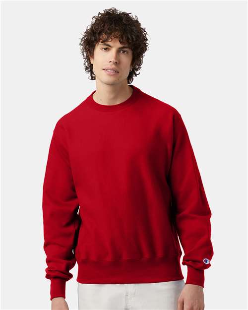 A person with curly brown hair wears a plain red Champion S149 sweatshirt and light-colored pants, standing against a plain light background.