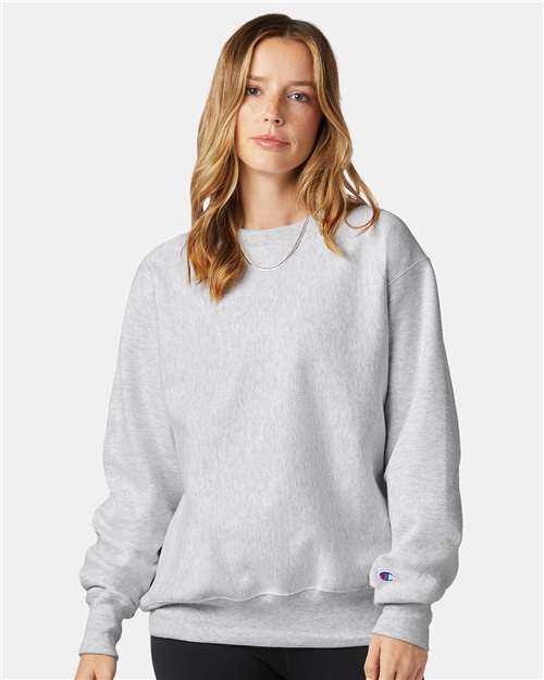 A woman with long, light brown hair wears the Champion S149 sweatshirt in light gray with black pants, standing against a plain light background. The iconic Champion logo is visible on the sleeve.