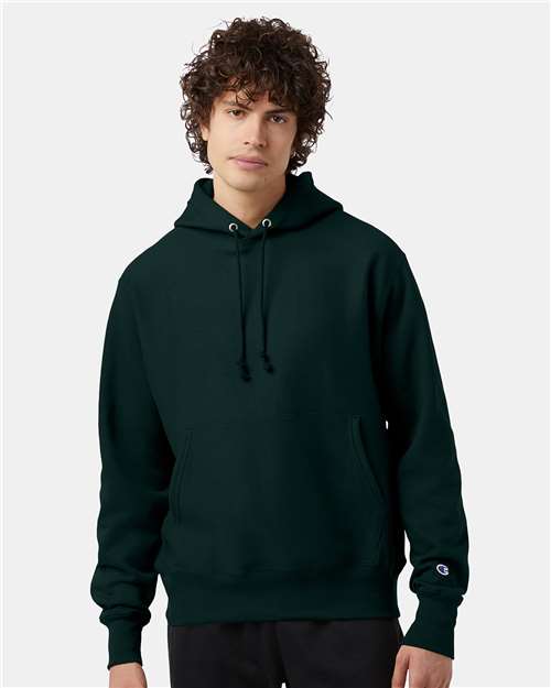A young person with curly hair wears a dark green Champion S101 hoodie with black drawstrings and a front pocket, standing against a plain light gray background.