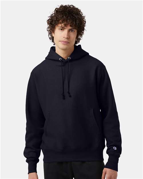 A young person with curly hair wears a Champion S101 plain black sweatshirt with a front pocket and drawstrings, standing against a light gray background.