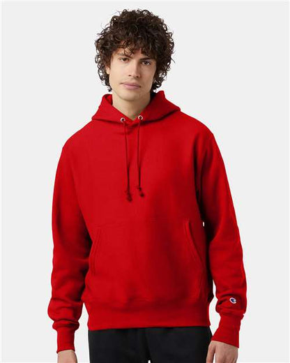 A young person with curly brown hair wears a bright red Champion S101 sweatshirt with front pockets and drawstrings, standing against a plain light gray background.