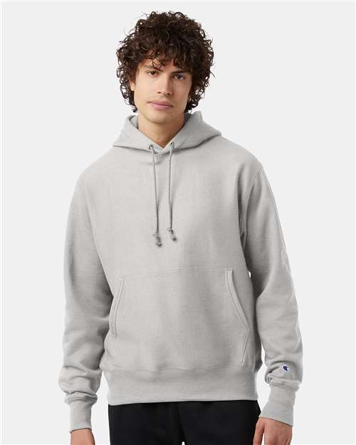 A young man with curly brown hair wears the Champion S101 hoodie, featuring a front pocket and drawstrings, against a plain light background.