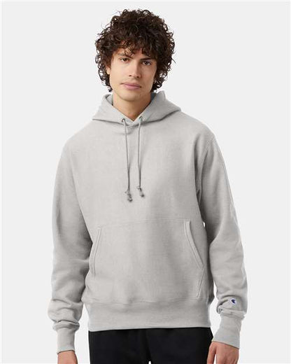 A young man with curly brown hair wears the Champion S101 hoodie, featuring a front pocket and drawstrings, against a plain light background.