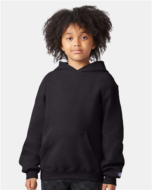 A young child with curly hair, wearing a Champion S790 plain black cotton-polyester hoodie with a front pocket, stands facing forward against a neutral background.