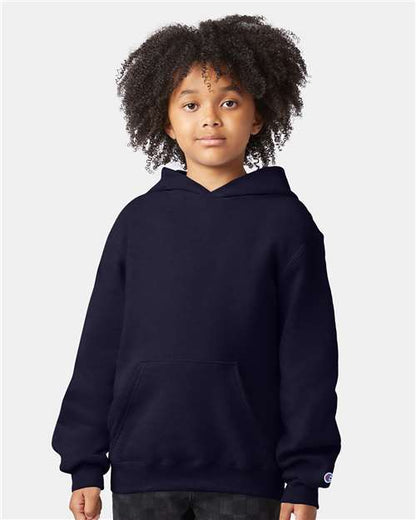 A young child with curly hair faces forward, wearing a Champion S790 dark navy cotton-polyester hoodie with a front pocket. The background is plain and light-colored.