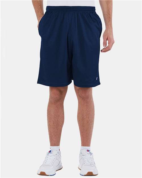 A person models Champion S162 navy blue athletic shorts, made from polyester mesh, paired with a white shirt and white sneakers. They stand against a plain white background, hands in pockets, shown from the waist down.