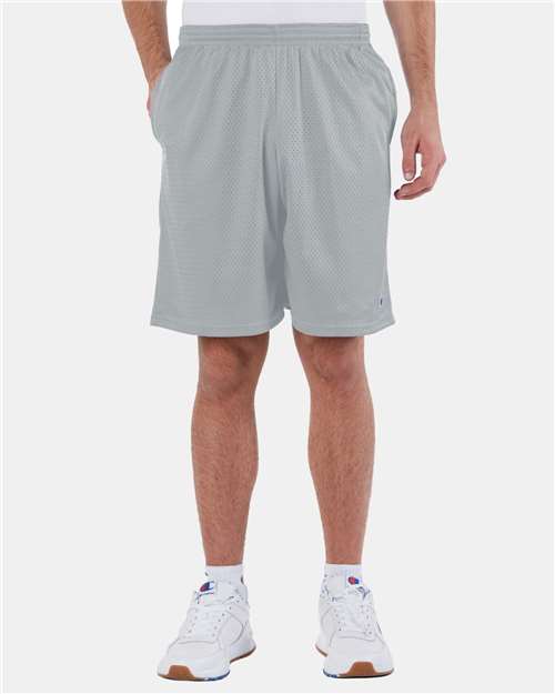 A person wears light gray Champion S162 polyester mesh athletic shorts, a white shirt, and white sneakers, standing against a plain white background with hands in the shorts’ pockets.