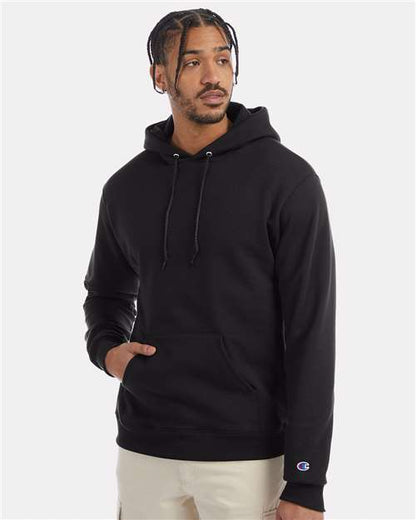 A man with braided hair wears the Champion S700 black hoodie made from recycled polyester, featuring a front pocket, and beige pants. He stands with one hand in his pocket, looking to the side against a plain background.