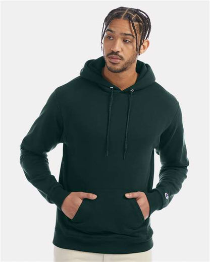 A man with braided hair wears a dark green Champion S700 hoodie made from recycled polyester, featuring front pockets, and looks slightly to the side against a plain light gray background.