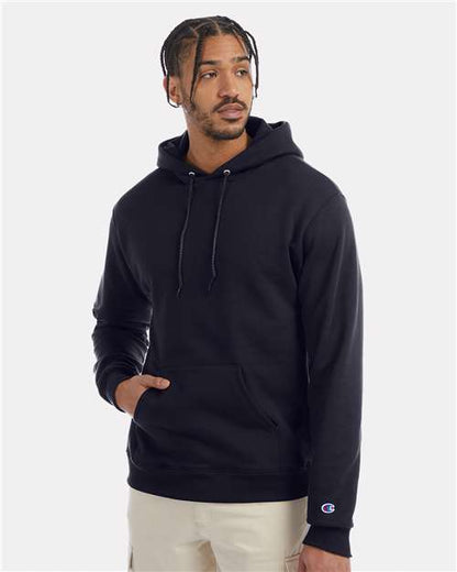 A man wearing a black Champion S700 hoodie with a front pocket and light-colored pants stands against a plain light background, looking slightly to the side.