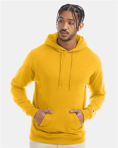 A man with braided hair wears a bright yellow Champion S700 hoodie, made from recycled polyester, hands in the front pocket, looking to the side against a simple light background.